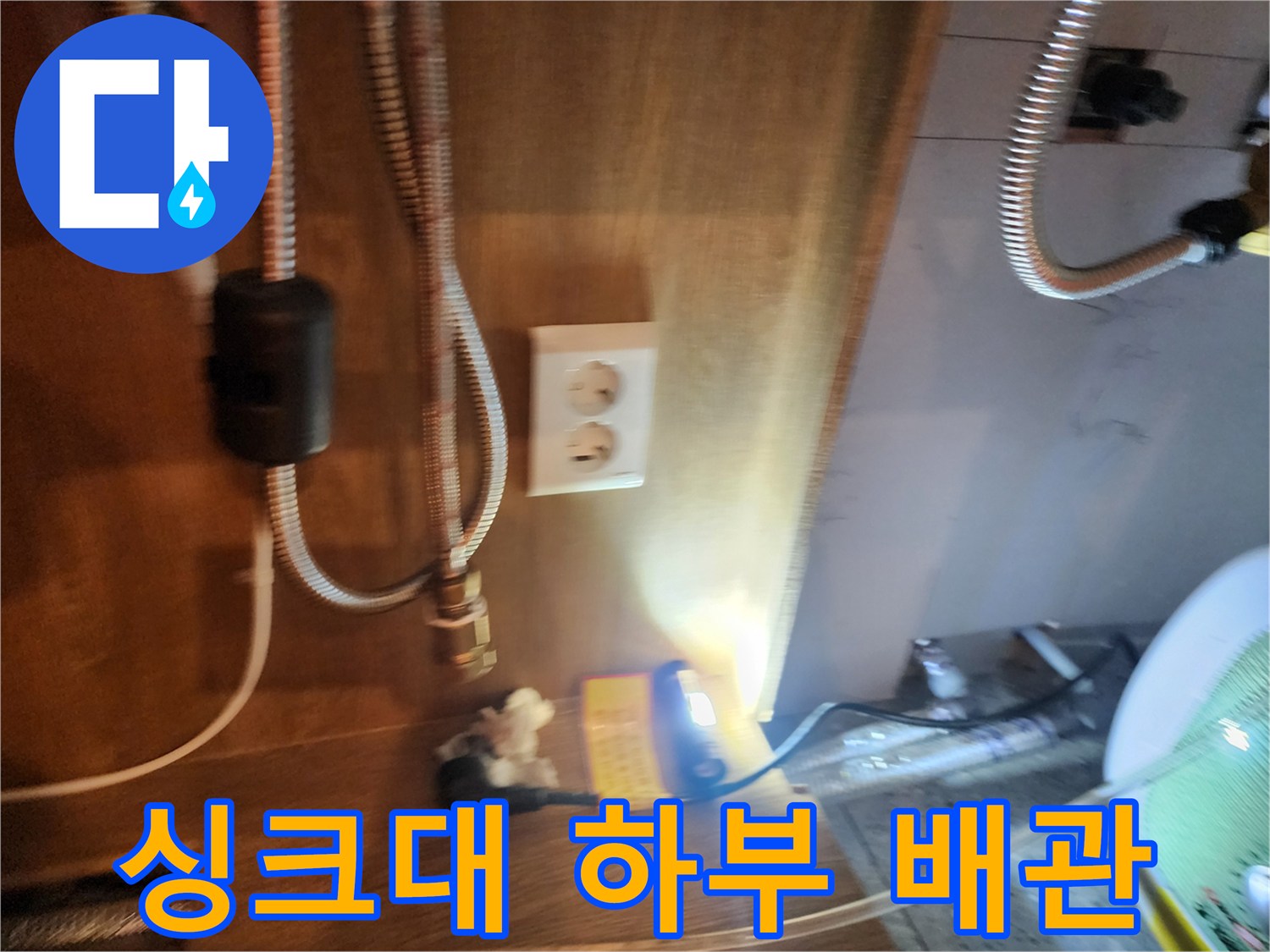 주방 싱크대 하부의 전기 온수기 연결 호스와 벽면 콘센트 주변 모습입니다.