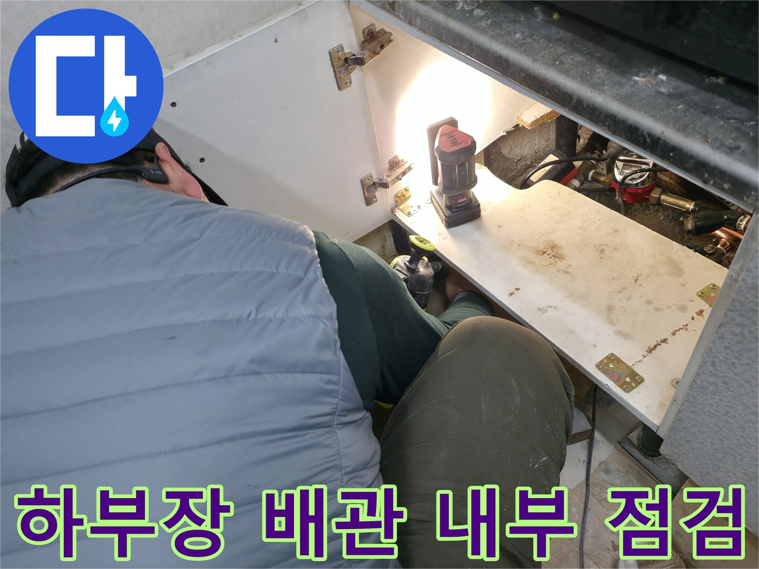 싱크대 아래쪽 어두운 공간의 배관을 조명으로 비추며 검사하는 장면