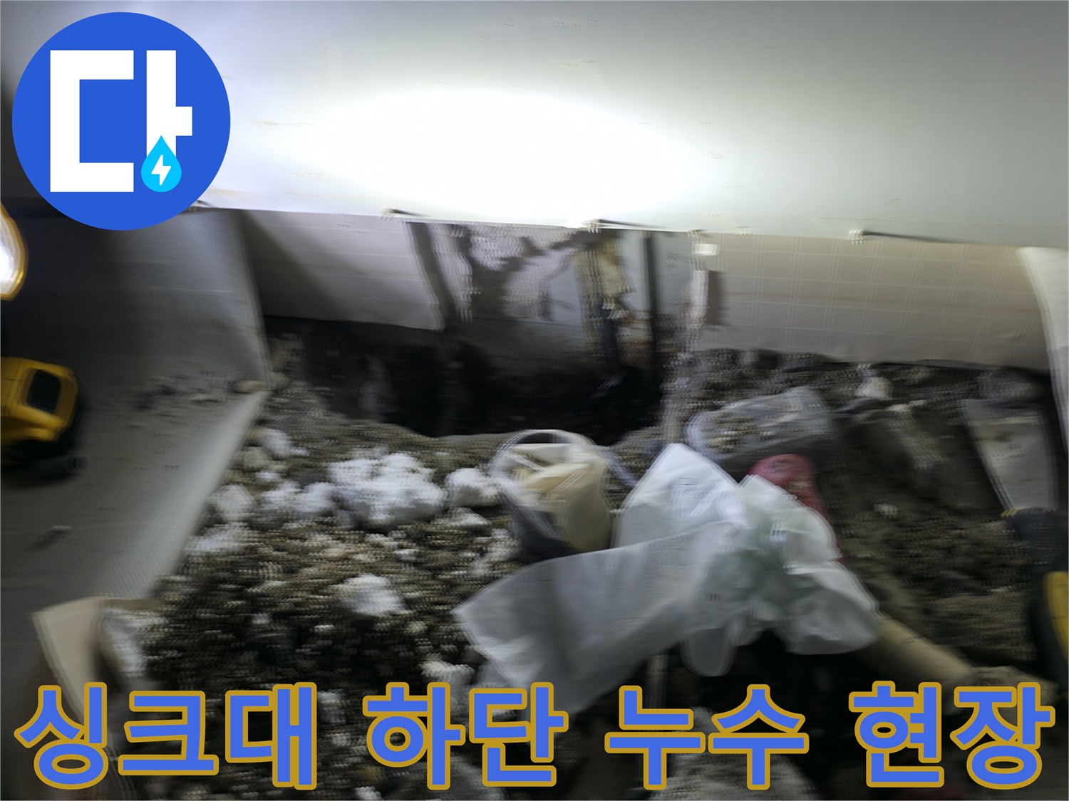 작업용 전등으로 비춘 싱크대 아래 젖은 바닥과 배관 모습입니다.