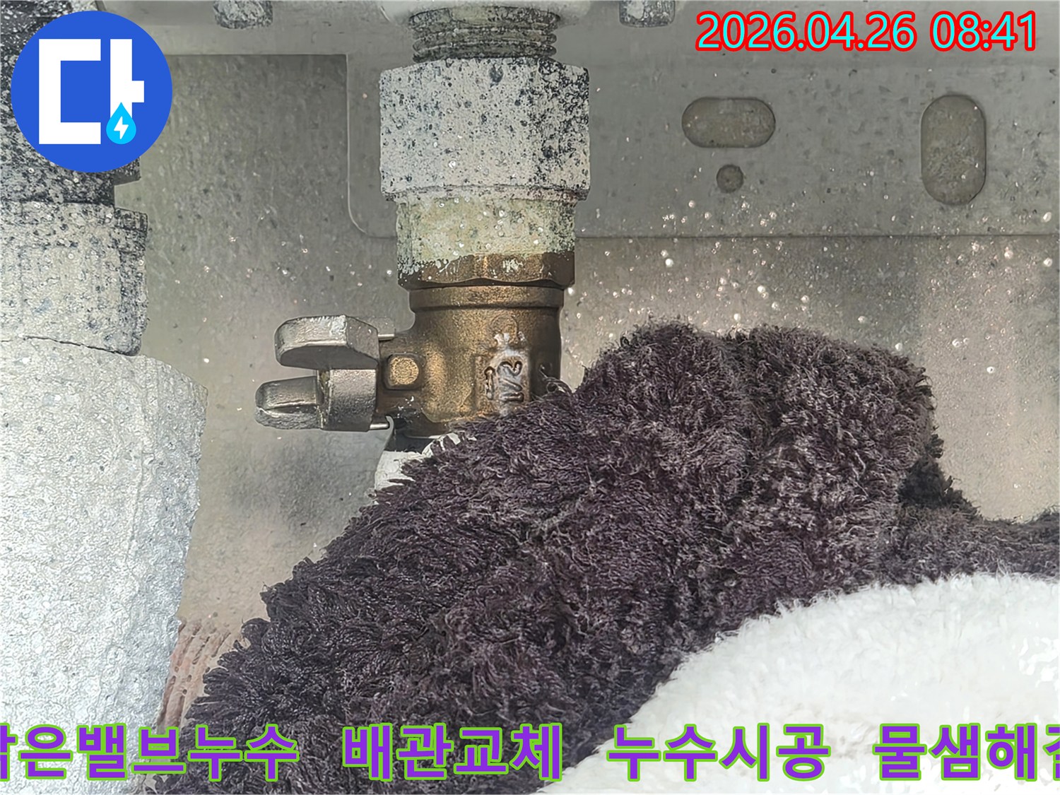 오래된 수도 밸브 주변에 하얀 침전물과 함께 물이 새는 모습-수건으로 물기를 흡수하고 있는 현장-노후된 배관의 누수 문제를 보여줍니다