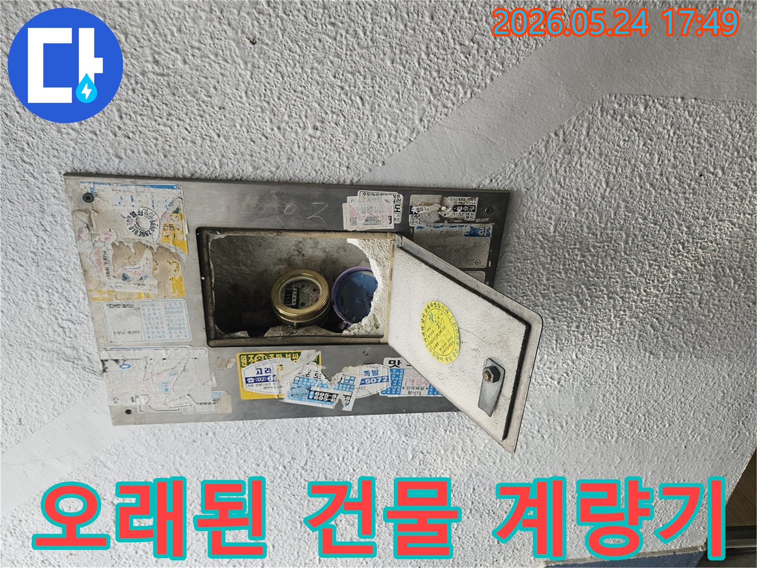 오래된 건물 외벽에 설치된-수도계량기함이 열려있고-내부 상태를 점검하는 모습입니다.