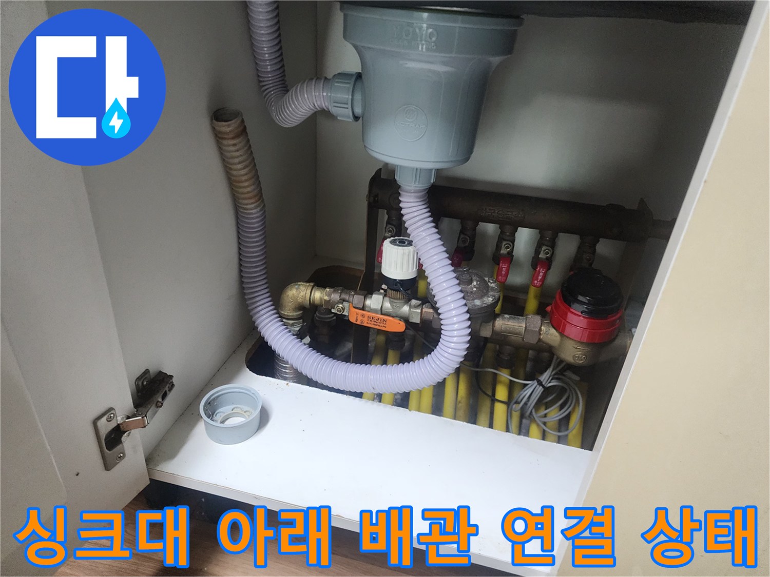 회색 트랩과 노란 밸브들이 모여있는 싱크대 하부장 내부 전경