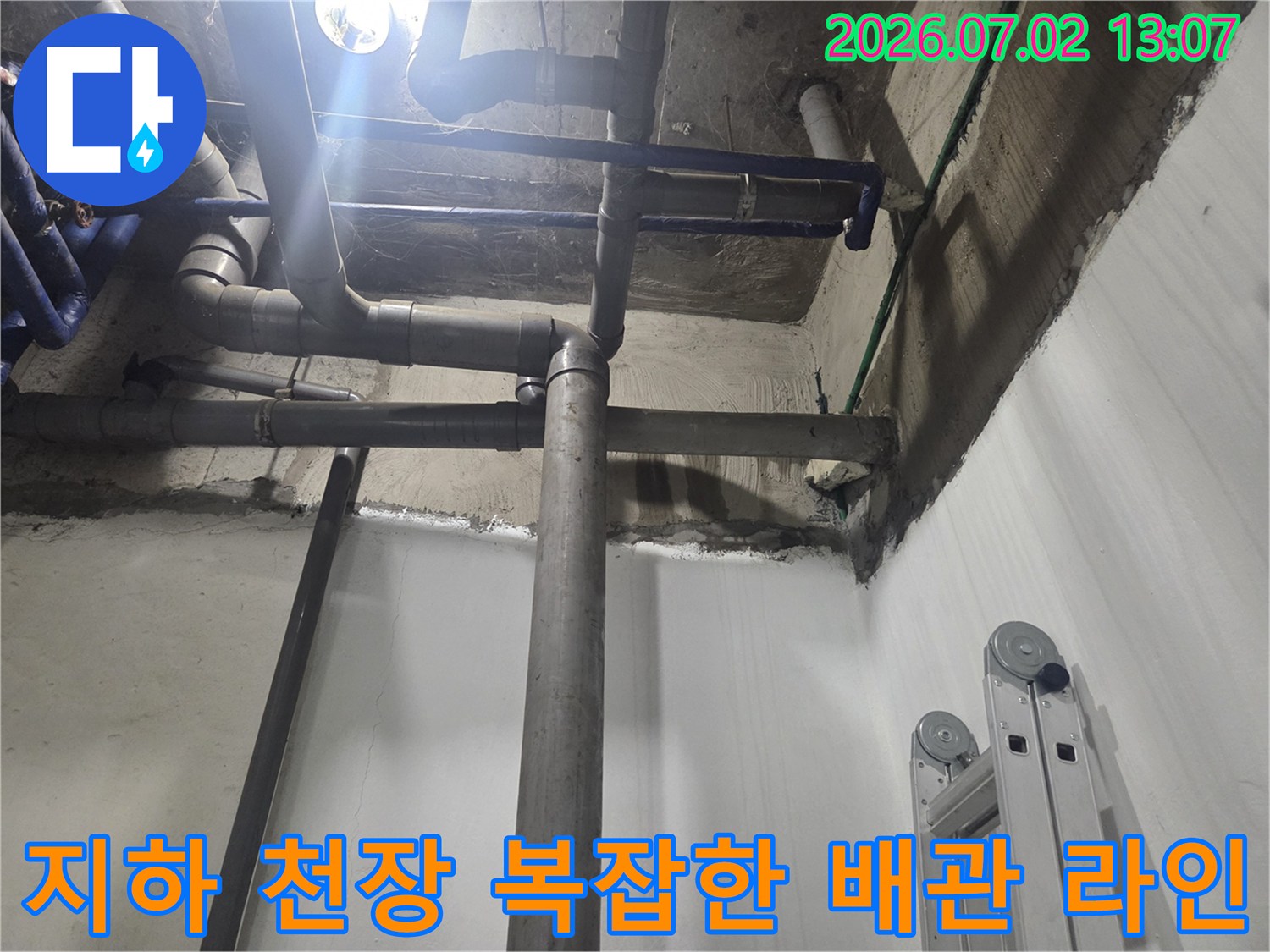지하 공간의 콘크리트 천장에 설치된 다양한 굵기의 회색 배관-라인과 파란색 보조 배관 모습