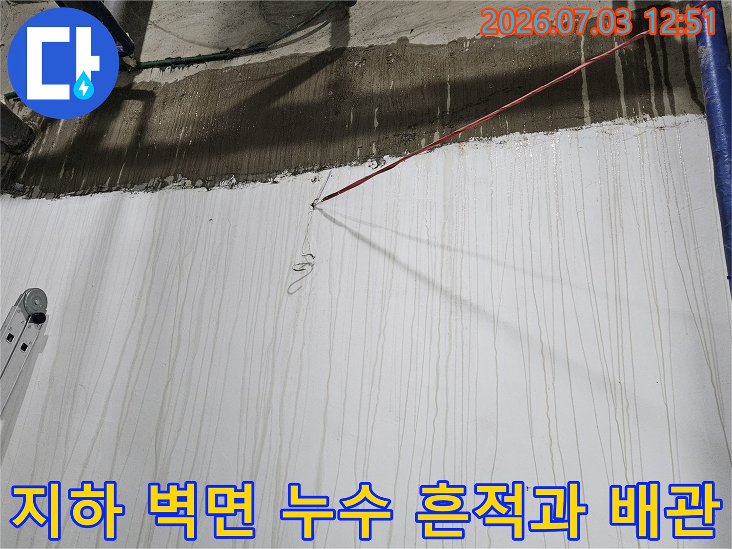 지하 공간의 흰색 벽면에 세로로 길게 형성된 누수 얼룩과 상단 콘크리트 층-파란색 보온재 배관 모습
