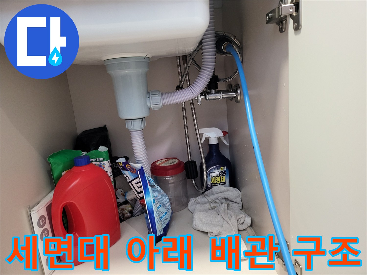 세면대 아래쪽의 복잡한 배관과 세제통 등이 놓여 있는 현장 모습입니다.