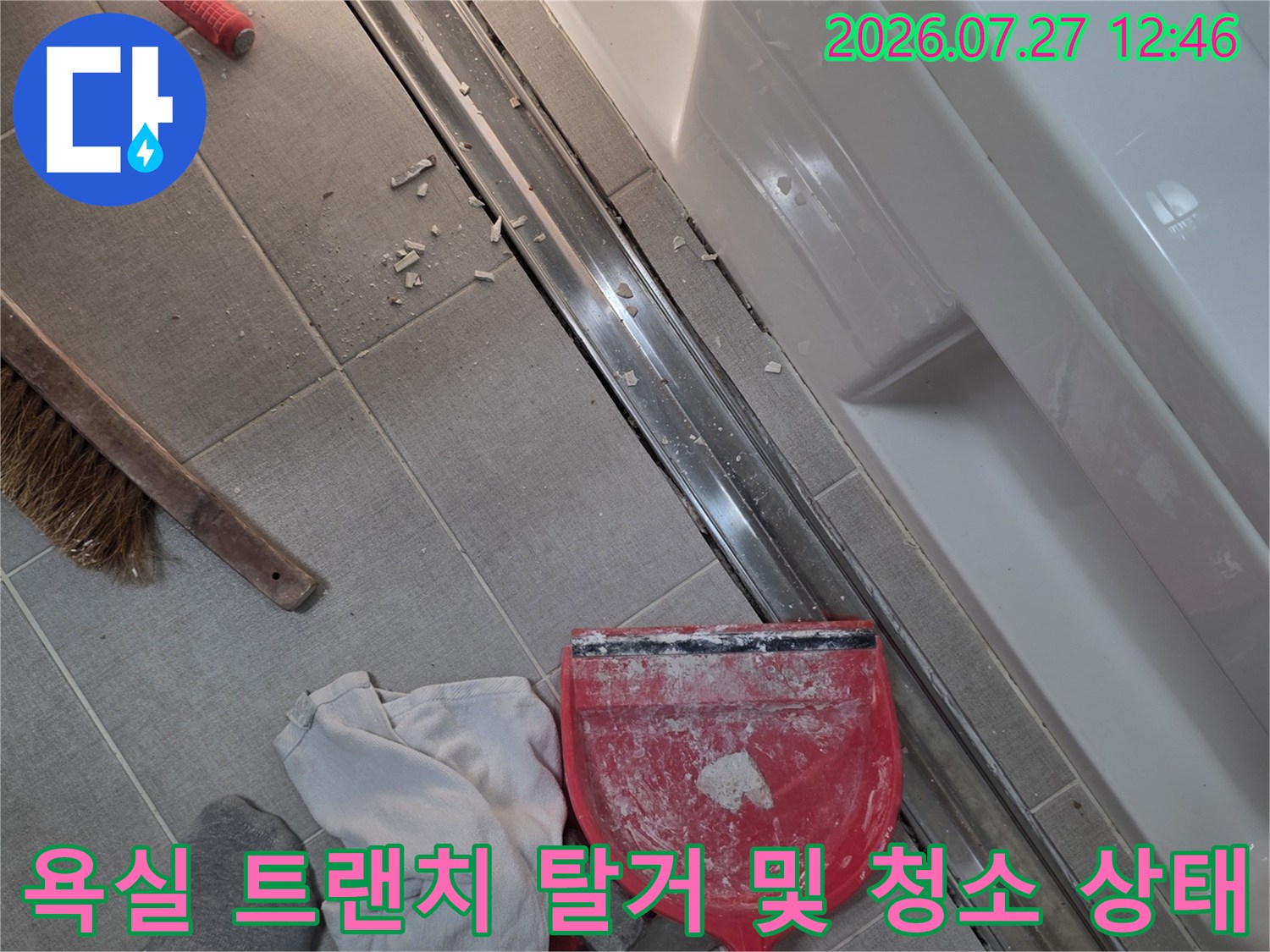 금속제 긴 배수구가 바닥에서 분리되어 옆으로 밀려나 있고 주변에 빨간 쓰레받기와 빗자루가 놓인 모습