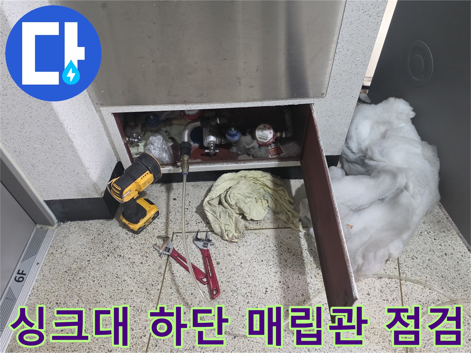 금속제 배관들이 연결된 싱크대 내부 공간과 바닥에 놓인 전동 드릴 사진
