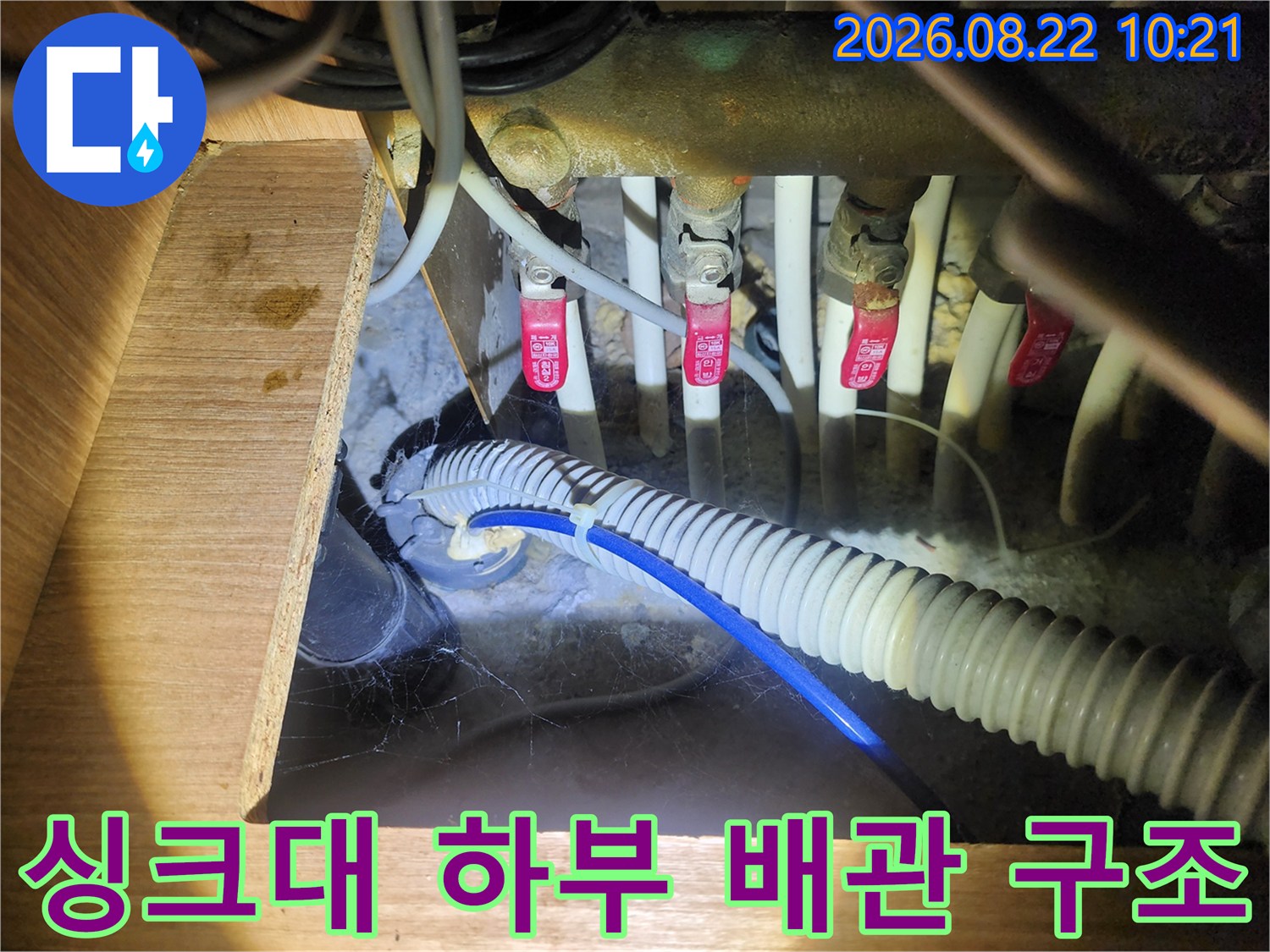 싱크대 밑에 설치된 다양한 수도 밸브와 주름진 배수 호스의 모습입니다.