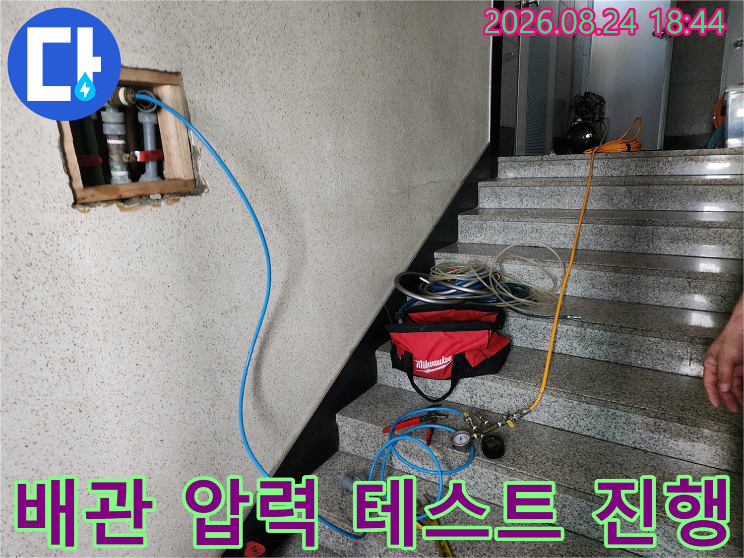 벽면 노출 배관에 연결된 파란색 호스와 압력 측정 장비 모습
