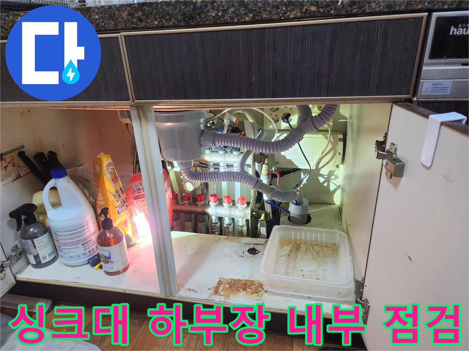 싱크대 하단 배관 연결 부위와 주변 세척 용품들이 놓여 있는 모습