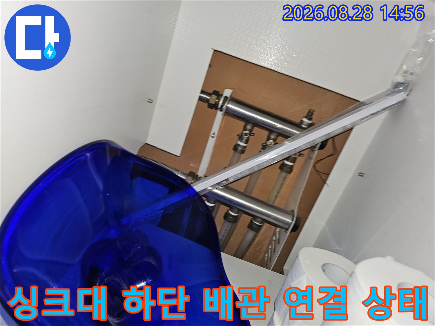싱크대 아래쪽 나무 받침 위에 설치된 금속 배관들의 연결 상태를 근접 촬영한 사진입니다.